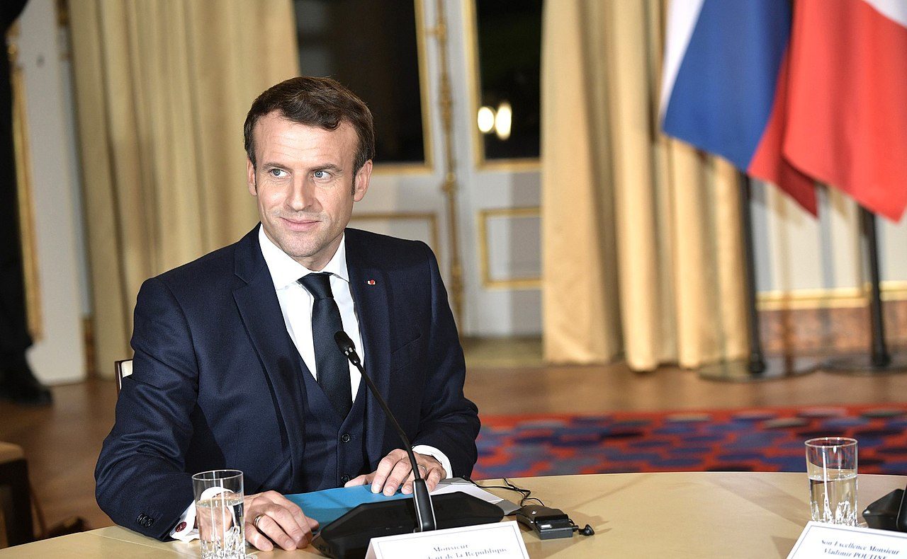 Emmanuel Macron est assis à un bureau dans une vaste salle de l'Elysée. Il regarde vers sa droite en souriant. Un petit micro, une feuille et un verre d'eau sont posés devant lui.
