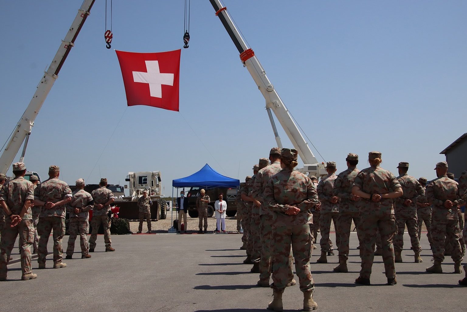 Des militaires debout de dos avec un drapeau de la Suisse suspendu dans les airs grâce à des grues