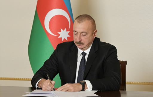 Le président de l'Azerbaïdjan Ilham Aliyev est assis à un bureau. Il est en train d'écrire sur un document. derrière lui se trouve un drapeau de l'Azerbaïdjan