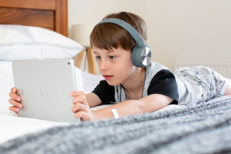 Un enfant allongé sur son lit regarde une tablette tactile avec un casque sur ses oreilles