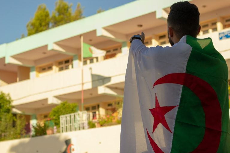 Un homme avec un drapeau algérien sur le dos pointe du doigt vers un autre étendard